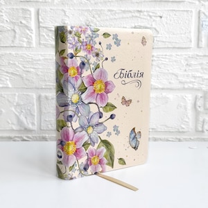 May include: A hardcover book with a floral design featuring pink, purple, and blue flowers, with the word "Бiблiя" on the cover. The cover also has butterfly illustrations. A wooden bookmark is inserted.