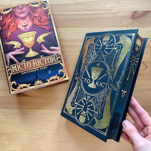 Könnte beinhalten: Zwei Hardcover-Bücher mit goldenen Akzenten und dem Titel "City of Ashes" in kyrillischer Schrift. Das Buch links zeigt eine Frau mit roten Haaren, die einen goldenen Kelch hält. Das Buch rechts hat einen dunkelblauen Einband mit goldenen Akzenten und einem Totenkopf- und Knochenkreuz-Design.