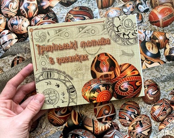 May include: A display of ornately decorated Easter eggs in shades of brown, black, and red. A book with Ukrainian text is held in the foreground, featuring similar egg designs. The eggs display geometric and animal motifs.