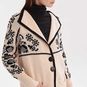 May include: A long, cream-colored cardigan with black floral patterns and trim. The cardigan has two large black buttons, two pockets, and a black collar. The sleeves are long and ribbed at the cuffs. The person in the image is wearing a black turtleneck.