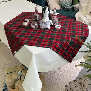 Pode incluir: Uma mesa festiva de Natal com um caminho de mesa xadrez vermelho e verde. A mesa é decorada com figuras de gnomos, enfeites e uma lanterna. Uma toalha de mesa branca e presentes embrulhados completam a cena.