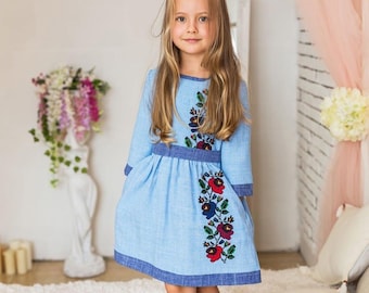 May include: A young girl wearing a light blue dress with embroidered flowers. The dress has a blue trim around the neckline and the bottom hem. The girl has long blonde hair and is standing in front of a white brick wall.