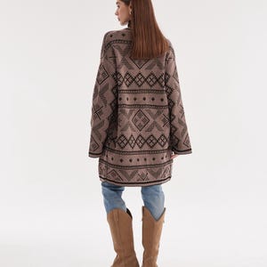 May include: A long-sleeved, tan cardigan featuring a repeating black geometric pattern of diamonds and horizontal lines. The garment is worn over light blue denim jeans and paired with tan suede knee-high boots with a pointed toe and block heel.