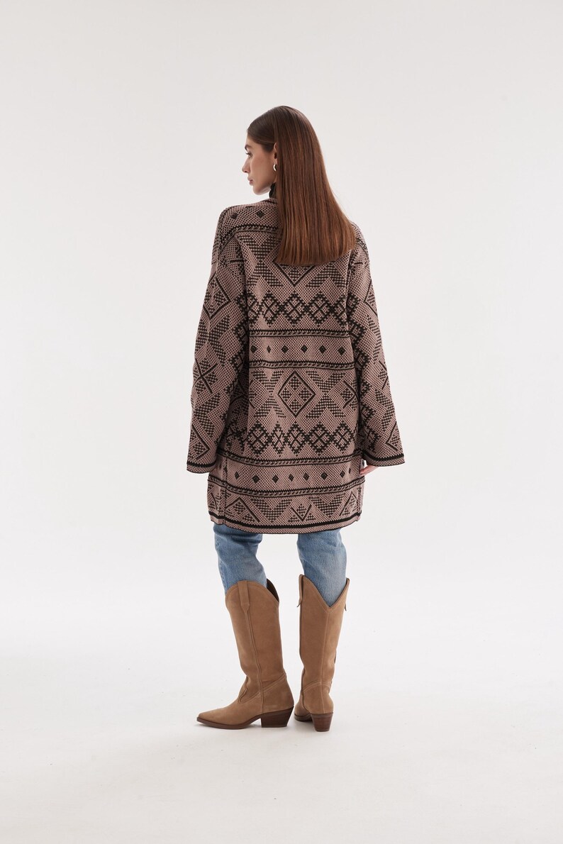 May include: A long-sleeved, tan cardigan featuring a repeating black geometric pattern of diamonds and horizontal lines. The garment is worn over light blue denim jeans and paired with tan suede knee-high boots with a pointed toe and block heel.