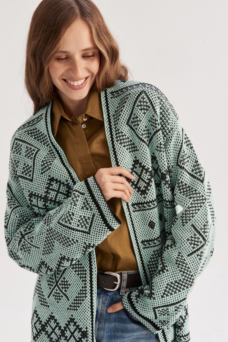 May include: A mint green and black open-front cardigan featuring a repeating geometric pattern throughout the knit fabric. The garment has long sleeves, a straight hem, and a solid black trim along the front opening and the sleeve cuffs.