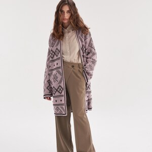 May include: A long-sleeved, open-front cardigan in light pink with a dark brown geometric pattern, worn over a beige button-down shirt and olive green wide-leg trousers with a high-waisted, overlapping front closure and dark brown shoes.