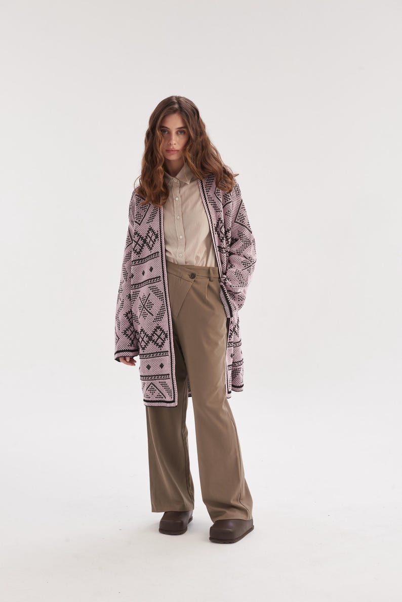 May include: A long-sleeved, open-front cardigan in light pink with a dark brown geometric pattern, worn over a beige button-down shirt and olive green wide-leg trousers with a high-waisted, overlapping front closure and dark brown shoes.
