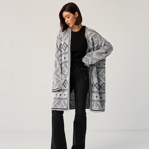 May include: A long, open-front cardigan featuring a repeating black and white geometric pattern. The garment has long sleeves and a straight hemline, worn over a black sleeveless top and black flared denim jeans with a dark wash finish.