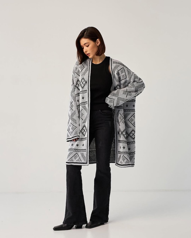 May include: A long, open-front cardigan featuring a repeating black and white geometric pattern. The garment has long sleeves and a straight hemline, worn over a black sleeveless top and black flared denim jeans with a dark wash finish.