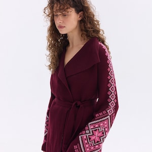 May include: A burgundy wrap cardigan with a shawl collar and a matching belt. The sleeves feature a geometric pattern in shades of pink and white. The cardigan is made of a soft, warm material.