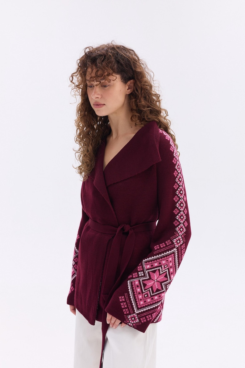 May include: A burgundy wrap cardigan with a shawl collar and a matching belt. The sleeves feature a geometric pattern in shades of pink and white. The cardigan is made of a soft, warm material.