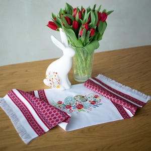 May include: A white ceramic bunny figurine with floral accents sits next to a vase of red tulips. A white table runner with red and white embroidered patterns and fringe is displayed on a wooden table. Easter decor.