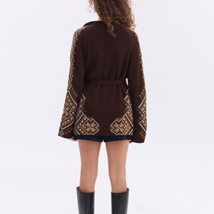 May include: A brown cardigan with intricate geometric patterns in beige and brown, featuring a matching belt. The sleeves and lower portion of the cardigan showcase detailed designs. The person is wearing black shorts and black knee-high boots.