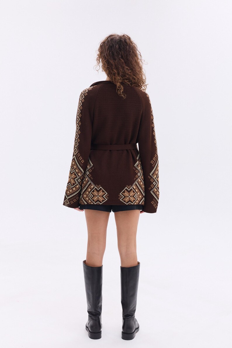 May include: A brown cardigan with intricate geometric patterns in beige and brown, featuring a matching belt. The sleeves and lower portion of the cardigan showcase detailed designs. The person is wearing black shorts and black knee-high boots.