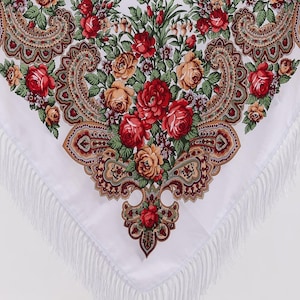 Op de afbeelding: Een witte driehoekige sjaal met een levendig bloemenpatroon van rode en oranje rozen, groene bladeren en ingewikkelde bruine paisley-motieven. De randen van de stof zijn afgewerkt met lange, dunne witte franjes langs de onderzijde.