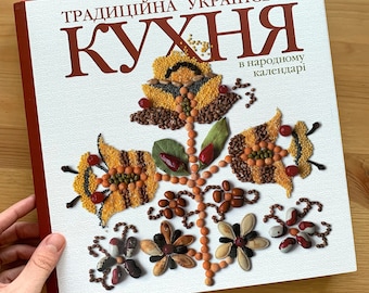May include: A hardcover cookbook titled "Традиційна Українська Кухня" with a floral design made of various grains and seeds. The cover features a red spine and text in Ukrainian.