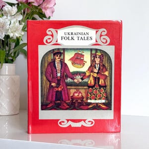 Pode incluir: Um livro de capa dura intitulado Ukrainian Folk Tales com uma capa vermelha brilhante. A frente apresenta uma ilustração central de duas figuras em trajes tradicionais de mãos dadas, com um pequeno navio acima e uma tigela de maçãs.