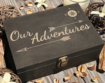 May include: A dark brown wooden box with a brass latch. The box has the words "Our Adventures" engraved on the top, along with a compass and two arrows.