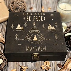 Pode incluir: Uma caixa de madeira preta com uma trava dourada e um desenho dourado de uma tenda indígena, montanhas e árvores. O texto "BE WILD, BE BRAVE BE FREE" e o nome "MATTHEW" estão gravados na caixa.
