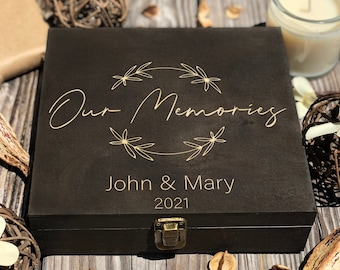 May include: A black wooden box with a gold latch. The box is engraved with the text "Our Memories" in a cursive font, surrounded by a wreath of leaves. Below the text is "John & Mary 2021".