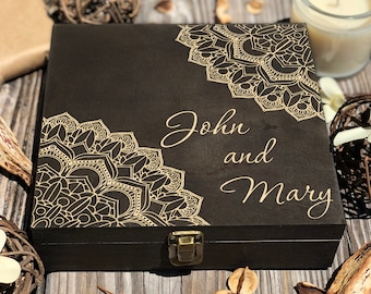 May include: A dark brown wooden box with gold mandala designs in the corners. The names "John and Mary" are scripted on the front. The box has a metal clasp and is on a wooden surface.