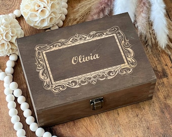 May include: A dark brown wooden box with a decorative frame and the name "Olivia" engraved on the lid. The box has a metal clasp and is likely used for storage or as a keepsake.