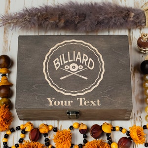 May include: A wooden box with a brown stain and a laser engraved design of a billiard ball and crossed cues. The text "BILLIARD" and "Your Text" are engraved on the box.