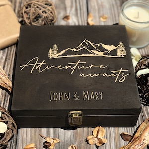 Puede incluir: Una caja de madera marrón oscuro con un diseño de montaña y árboles, y las palabras "Adventure awaits" en escritura cursiva. Los nombres "JOHN & MARY" están debajo. La caja tiene un cierre de metal.