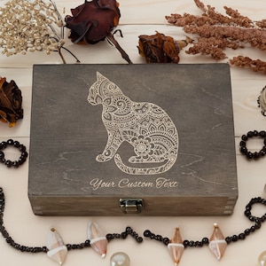 May include: A dark brown wooden box with a hinged lid and a latch. The box has a decorative design of a cat with intricate patterns engraved on the front. The text "Your Custom Text" is engraved below the cat design.