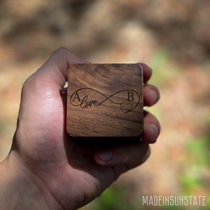 May include: A square wooden box with a dark wood grain. The top is engraved with the letters "A" and "B" on either side of an infinity symbol with the word "love" in cursive. A heart is next to the letter "B".