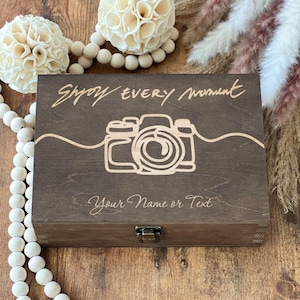 以下が含まれることがあります： 蓋が蝶番で開閉する、濃い茶色の木製の箱。金属製の留め金が付いている。箱には「Shoot Every Moment」という文字と、スタイリッシュなカメラの画像が刻まれている。「Your Name or Text」という文字も箱に刻まれている。