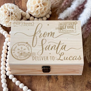 Könnte beinhalten: Eine Holzkiste mit einem Scharnierdeckel, verziert mit goldenen Lettern, die "von Santa, liefern an: Lucas" lauten. Die Kiste hat auch einen goldenen Stempel, der "Lieber Weihnachtsmann, Nordpol" lautet, und einen weiteren, der "Nicht vor Weihnachten öffnen" lautet.