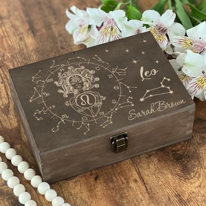 May include: A dark brown wooden box with a hinged lid and a latch. The box is decorated with an engraved design of a woman with a lion's head, surrounded by stars and zodiac symbols. The text "Leo" and "Sarah Brown" is engraved on the box.