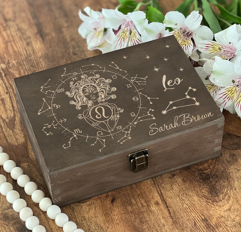 May include: A dark brown wooden box with a hinged lid and a latch. The box is decorated with an engraved design of a woman with a lion's head, surrounded by stars and zodiac symbols. The text "Leo" and "Sarah Brown" is engraved on the box.
