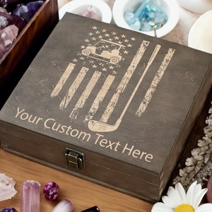 Pode incluir: Uma caixa de madeira com um design de bandeira americana desbotada, um carrinho de golfe e um taco de golfe. A caixa tem uma tampa com dobradiças e uma trava de metal. O texto "Your Custom Text Here" está gravado na frente da caixa.