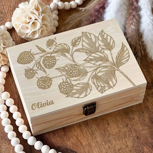May include: A wooden box with a hinged lid and a brass latch. The box is decorated with a laser-engraved design of a raspberry branch with leaves and berries. The name "Olivia" is engraved on the front of the box.