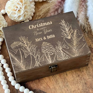 May include: A dark brown wooden box with a hinged lid and a brass latch. The box is decorated with a laser-engraved design of stylised leaves and flowers. The text "merry Christmas & happy New Year Alex & Julia" is engraved on the lid.