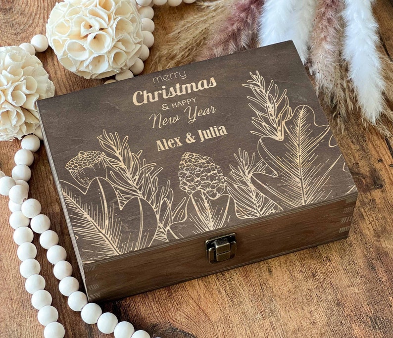 May include: A dark brown wooden box with a hinged lid and a brass latch. The box is decorated with a laser-engraved design of stylised leaves and flowers. The text "merry Christmas & happy New Year Alex & Julia" is engraved on the lid.
