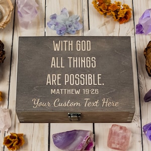 Puede incluir: Una caja de madera con tapa abatible y cierre. La caja está grabada con el texto "Con Dios todo es posible. Mateo 19:26" y "Tu texto personalizado aquí".