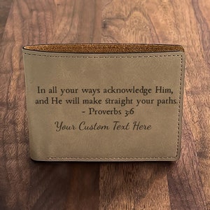 Puede incluir: Una cartera de cuero marrón con una cita de Proverbios 3:6, "Reconoce a Dios en todos tus caminos, y él allanará tus sendas." La cartera también tiene el texto "Tu texto personalizado aquí" grabado.