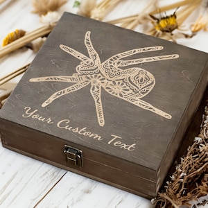 Pode incluir: Uma caixa de madeira castanho-escuro com tampa articulada e fecho de metal. A caixa é decorada com um desenho de aranha de estilo mandala dourado. O texto "Your Custom Text" está gravado na frente da caixa.