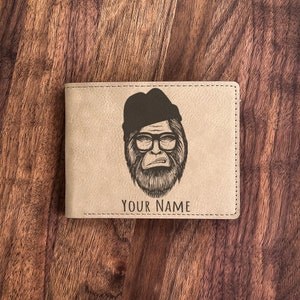 May include: Tan bi-fold wallet with a black illustration of a Sasquatch wearing a hat and glasses. The text "YOUR NAME" is below the illustration. The wallet is on a wooden surface.