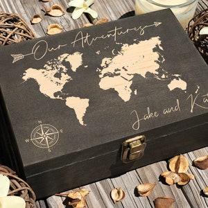 May include: A dark wooden box with a world map design and the words "Our Adventures" and "Jake and Kat" engraved on the lid. The box has a brass clasp and a compass rose design. The box is surrounded by decorative items.