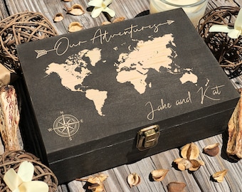 May include: A dark wooden box featuring a world map design, with the words "Our Adventures" and "Jake and Kat" engraved on the lid. The box has a brass clasp and a compass rose design. Decorative items surround the box.
