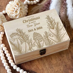 May include: A wooden box with a hinged lid, decorated with a gold-coloured design of leaves and branches. The box has a gold-coloured inscription that reads "merry Christmas & happy New Year Alex & Julia".