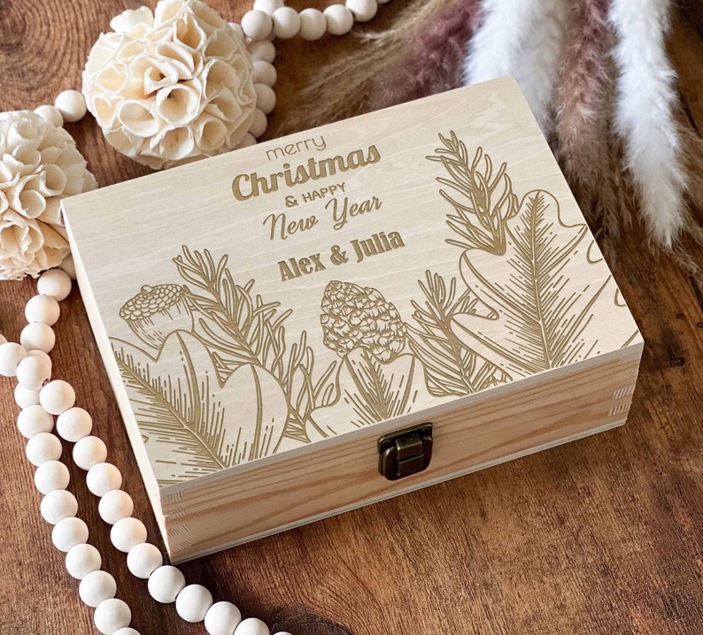 May include: A wooden box with a hinged lid, decorated with a gold-coloured design of leaves and branches. The box has a gold-coloured inscription that reads "merry Christmas & happy New Year Alex & Julia".