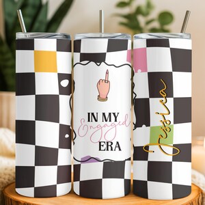 May include: A cylindrical tumbler with a black and white checkered pattern, a metal straw, and a lid. The design features a hand with a ring, the text "IN MY Engaged ERA," and the name "Jessica" in gold script over a green square accent.