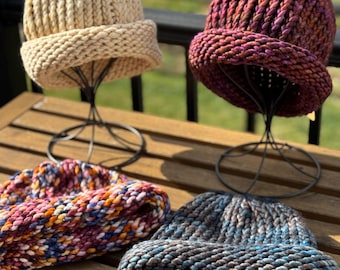 May include: Four chunky knit beanies in various colors are displayed on a wooden table. The beanies include beige, burgundy, and a multicolored option with blue, orange, and pink. The hats are displayed on black metal stands.