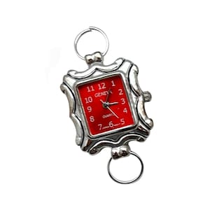 以下が含まれることがあります： 赤い文字盤と正方形のフレームを持つ銀色の腕時計。時計の文字盤には黒い針と黒い数字があります。時計は留め具付きの銀色のバンドが付いています。