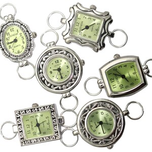 May include: Six silver-tone pocket watches with green faces and black hands. The watches are connected by silver-tone chains. Each watch has a different design around the face.